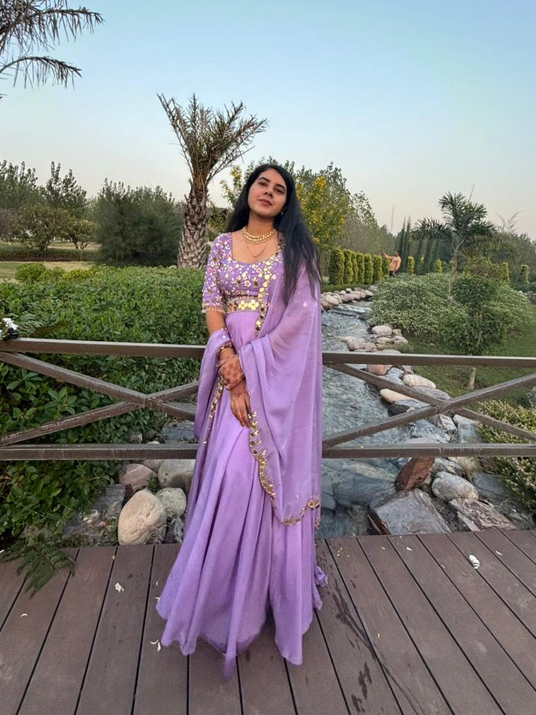 A woman wearing a lavender purple traditional dress with gold embroidery stands on a wooden deck in a landscaped outdoor garden with palm trees and a small stream.